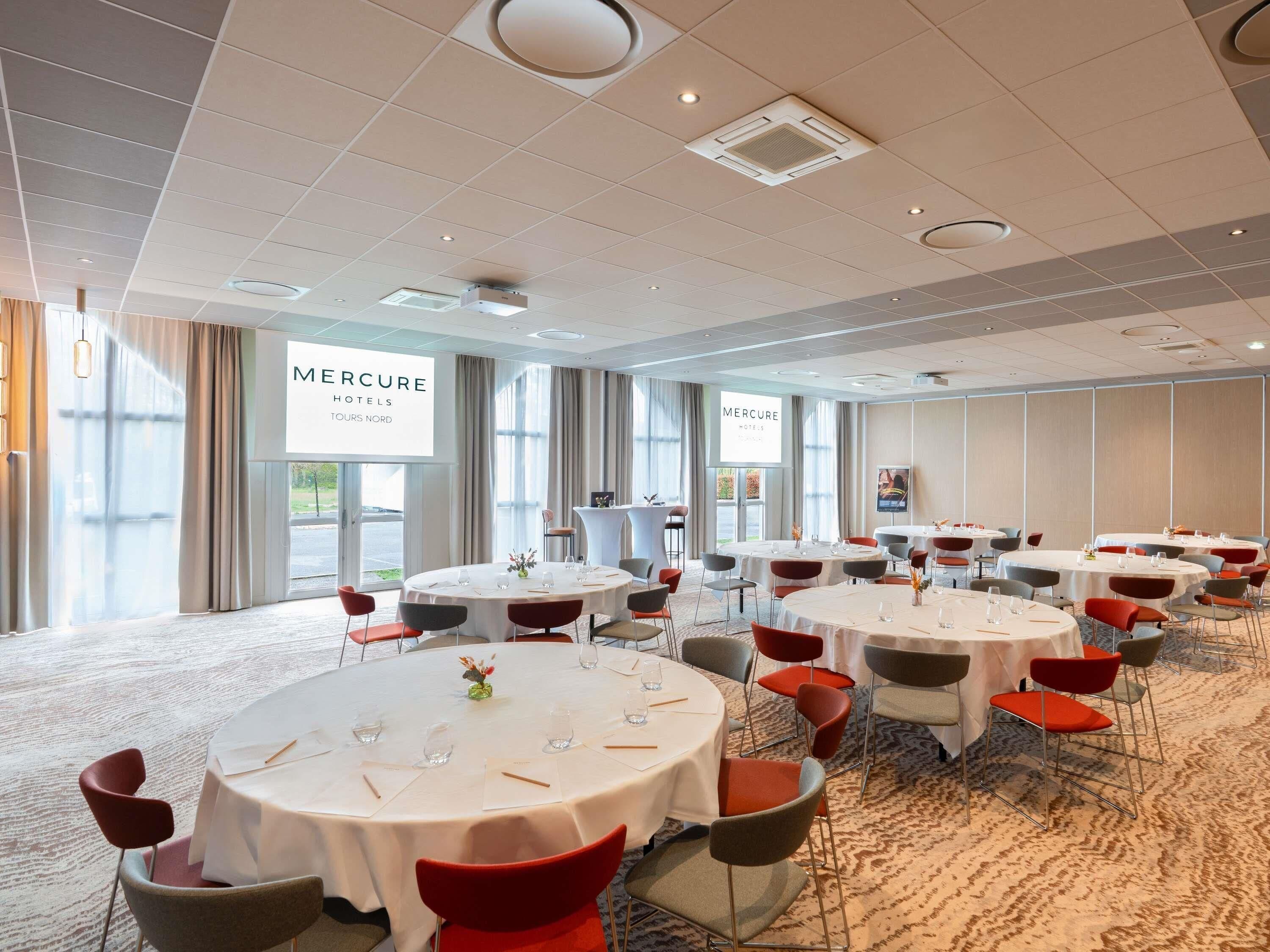 Un salón de conferencias con mesas redondas, sillas y una gran pantalla que muestra 'Mercure Hotels Tours Nord'.