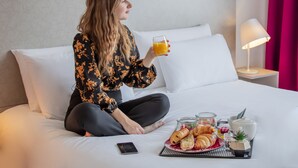 Una persona sentada en una cama con artículos de desayuno, como cruasanes, un vaso de jugo de naranja y una planta pequeña.