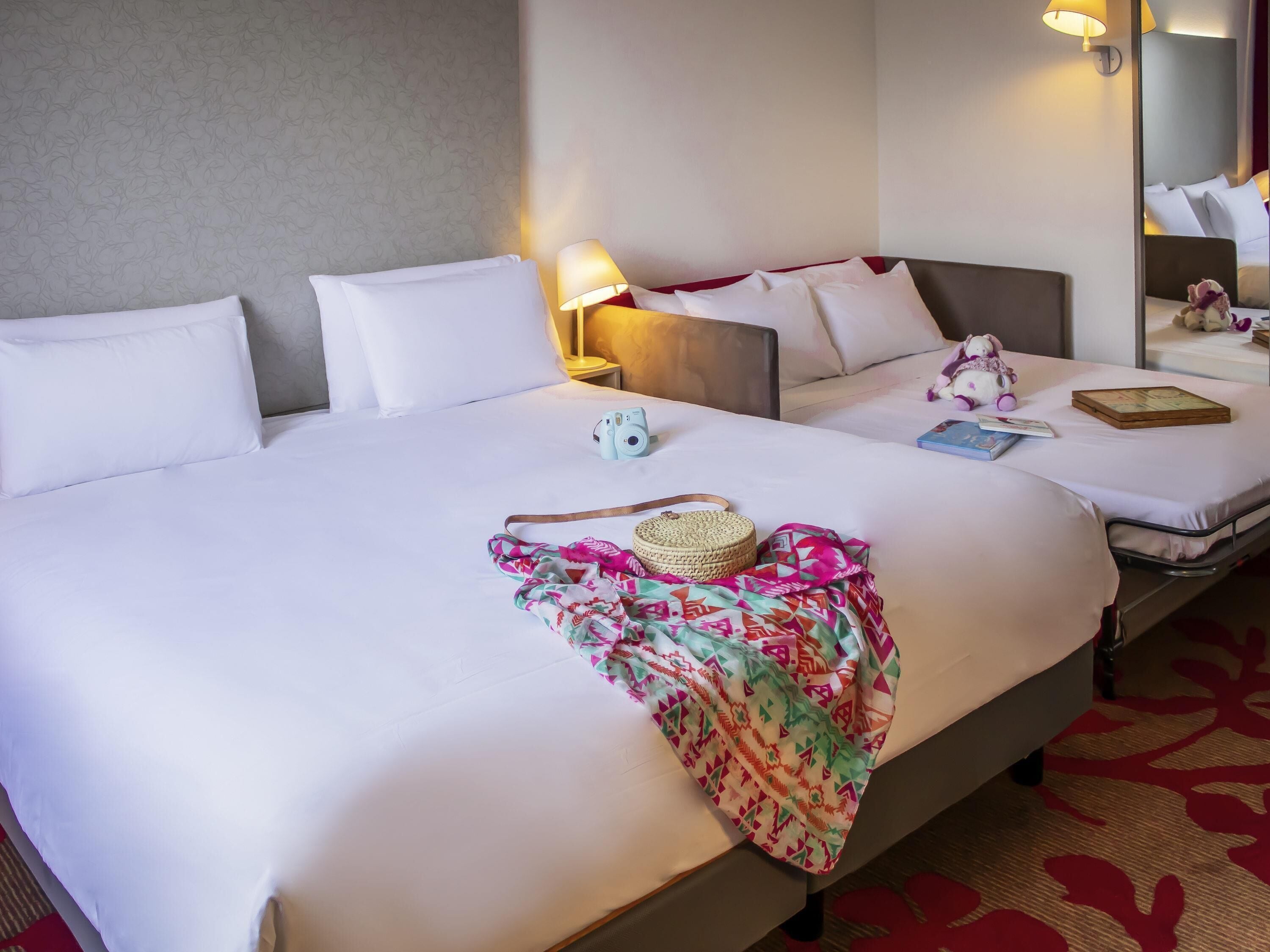 Habitación de hotel con dos camas, una lámpara de noche, un espejo, un peluche y un libro.