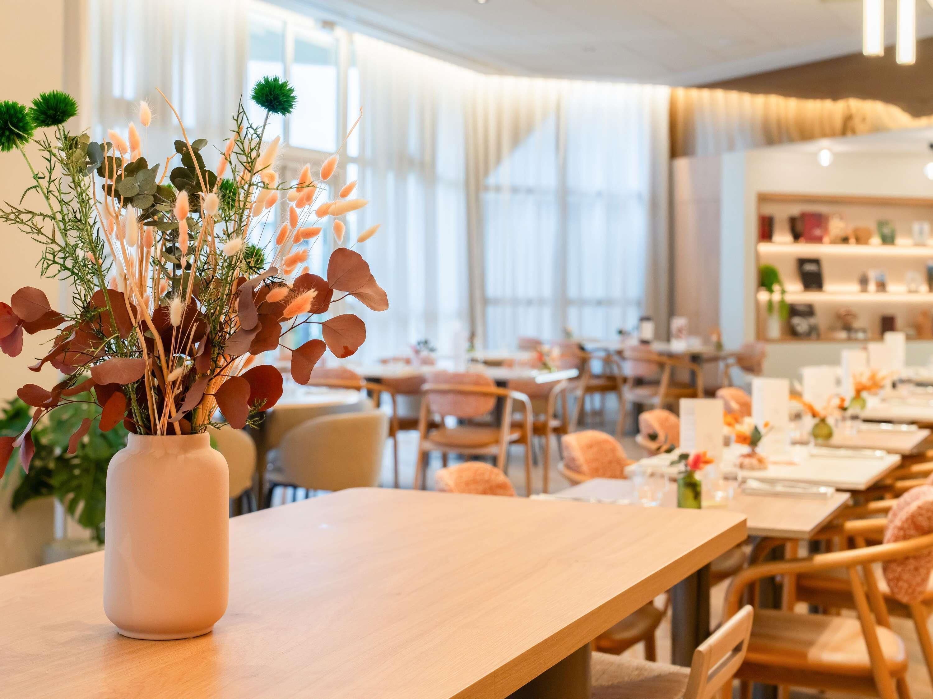 Een restaurant met een houten tafel en stoelen, een vaas met bloemen, en een licht interieur.