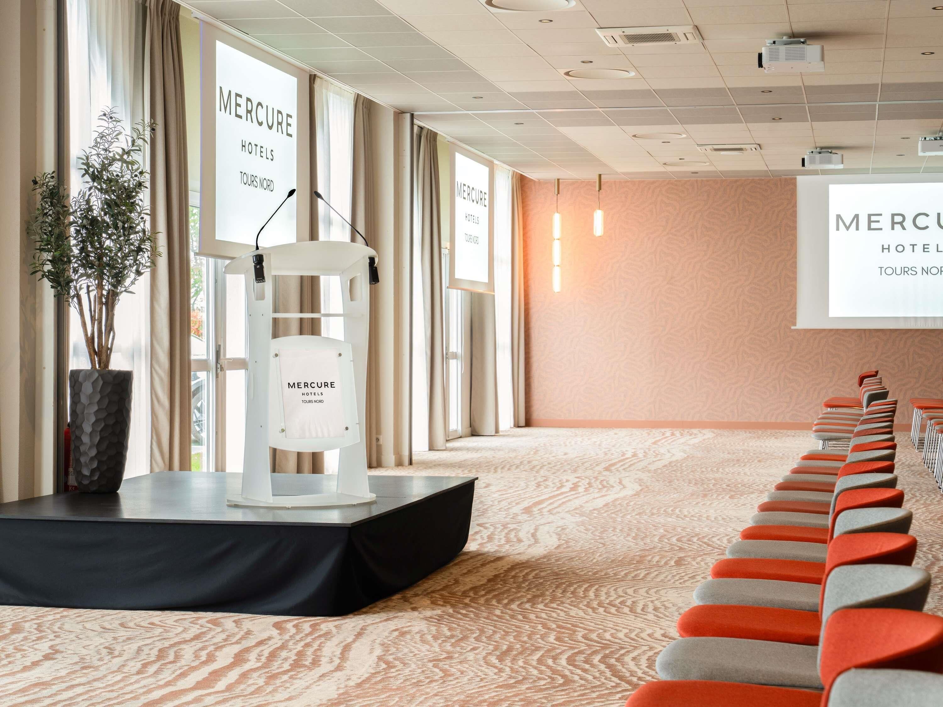 Een vergaderzaal met een spreekgestoelte, rijen stoelen en grote ramen.