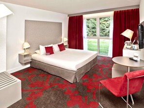 A hotel room with a bed, two bedside lamps, a TV, a small table, and a red chair.