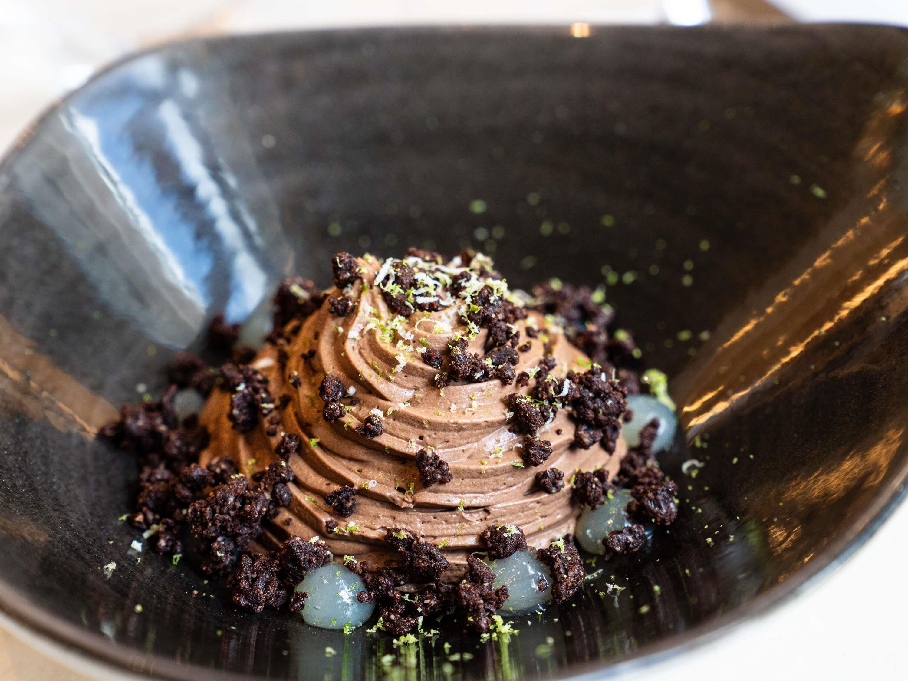 A dessert with chocolate mousse and crumbled chocolate pieces, garnished with green specks, served in a dark bowl.