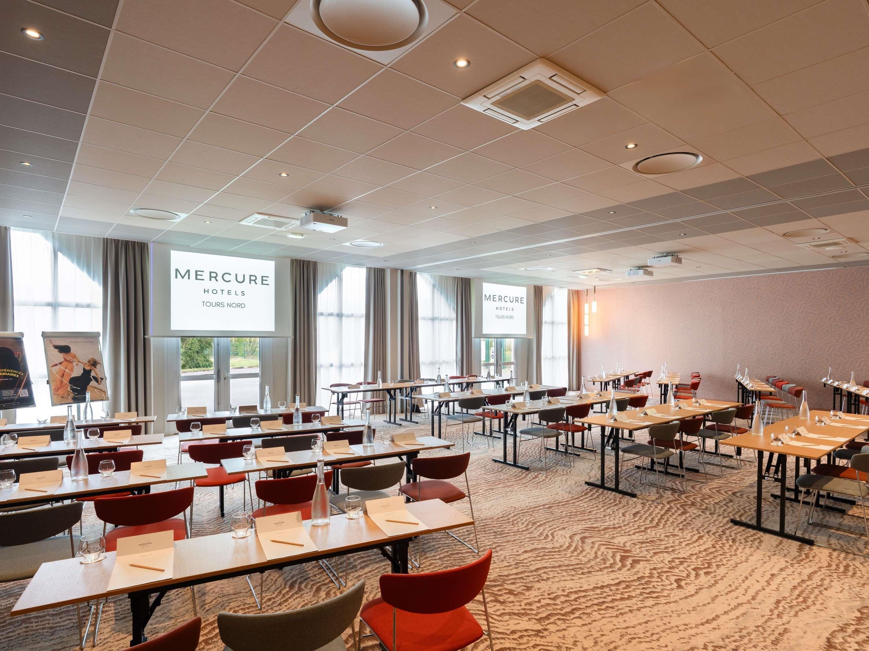 Ein Konferenzraum mit mehreren Tischen und Stühlen, Namensschildern und einem großen Bildschirm, der 'Mercure Hotels Tours Nord' anzeigt.