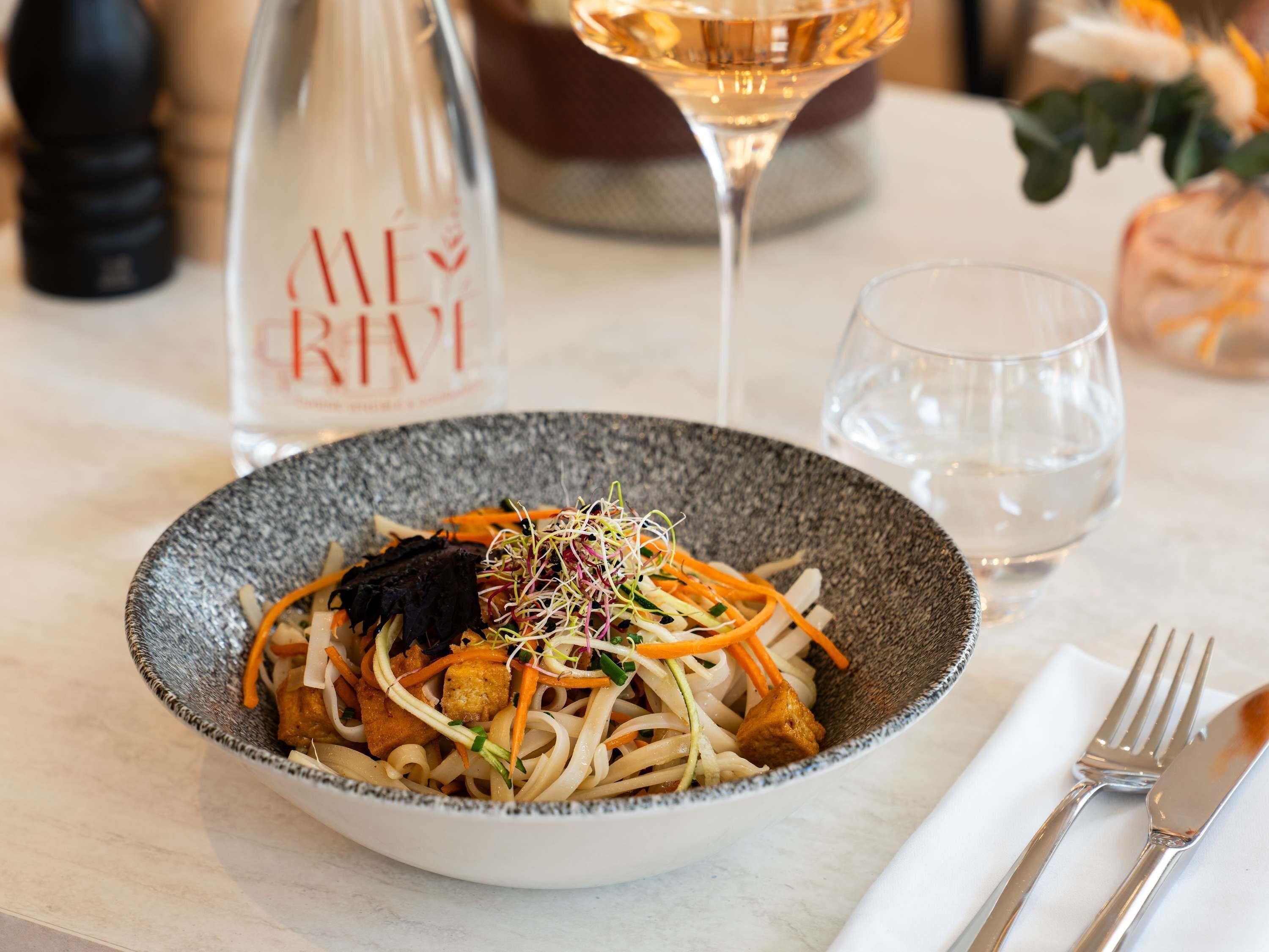 A bowl of noodle dish with vegetables and sprouts, a wine glass, and a water glass on a table.