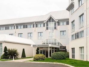 Un hotel moderno a più piani, con un ingresso centrale caratterizzato da vetrate, numerose finestre e un giardino curato con diverse piante.