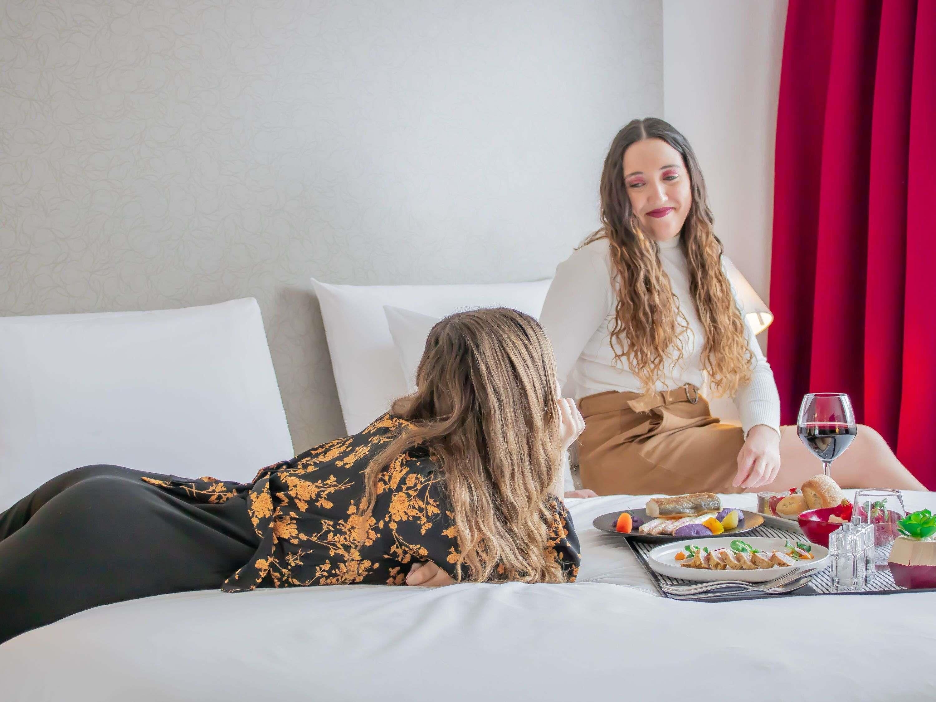 A hotel room with a bed, pillows, a dining table with plates of food, glasses, and a wine glass.