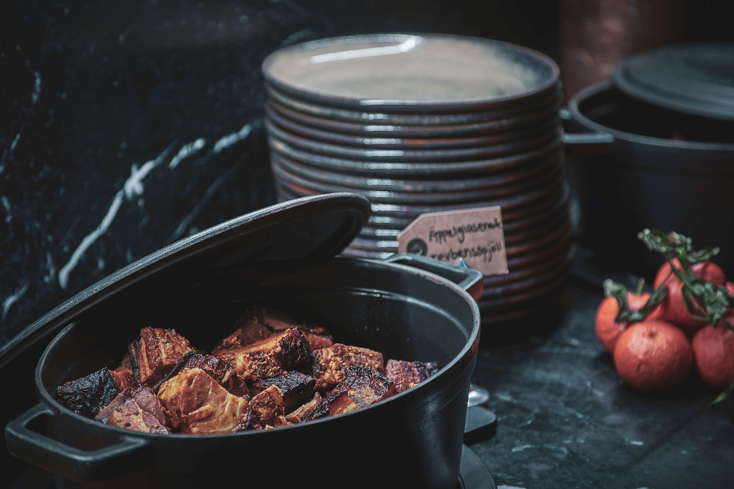 A pot of cooked meat, stacked plates, and fresh tomatoes on a dark surface.