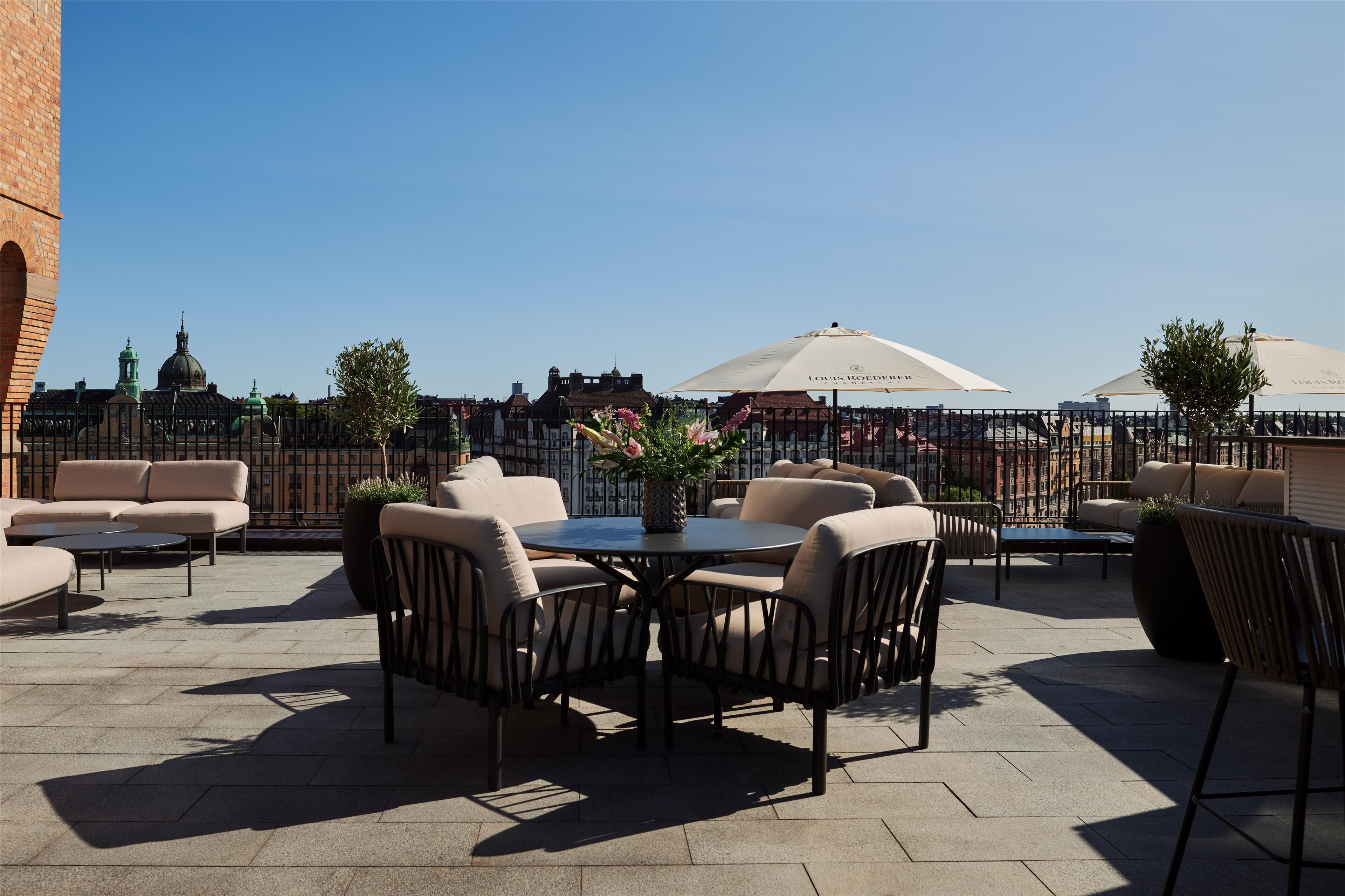 An outdoor seating area with round tables and chairs, a large umbrella, and a view of historic buildings.