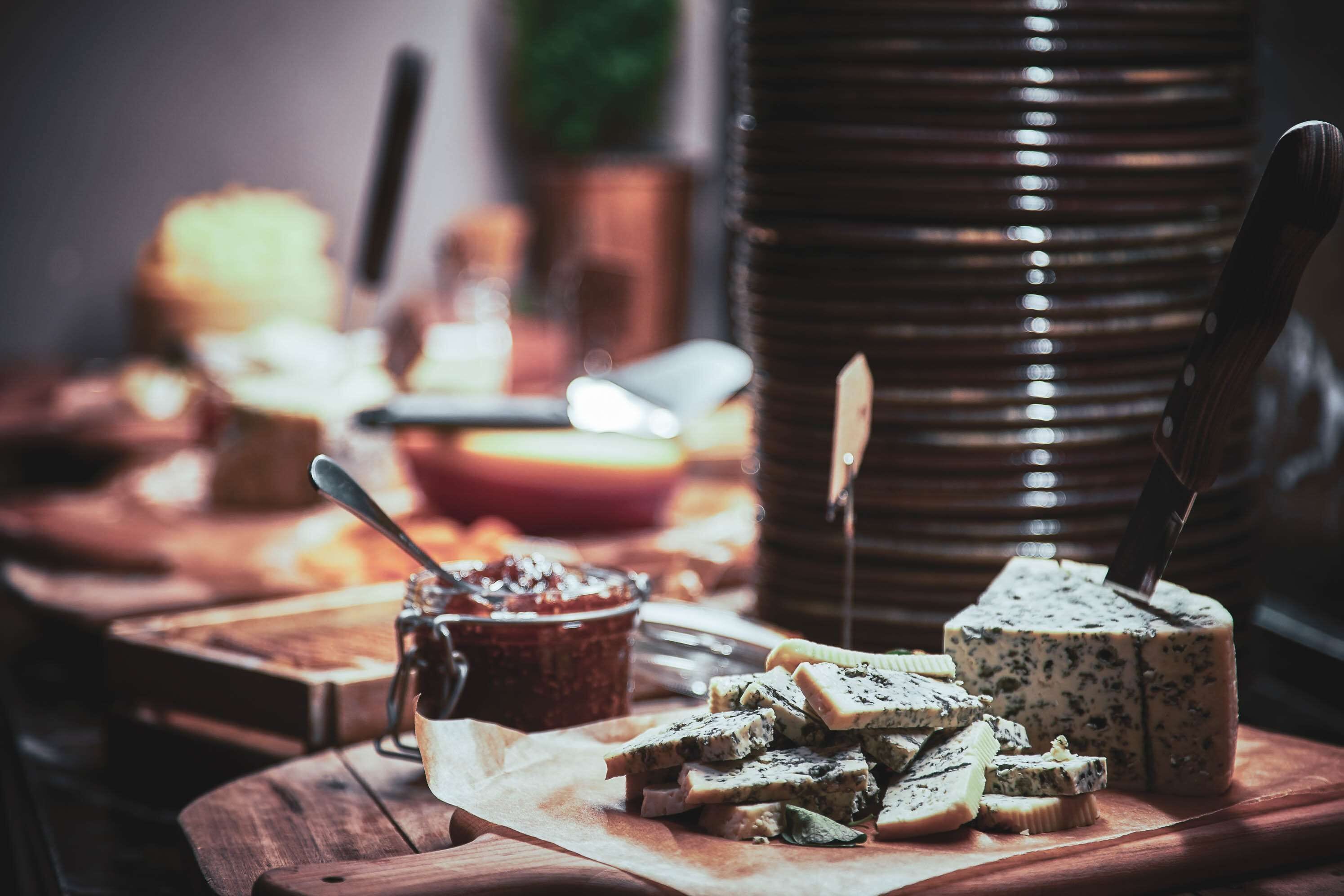Uma mesa de madeira com queijos variados, uma faca e um pote de geleia.