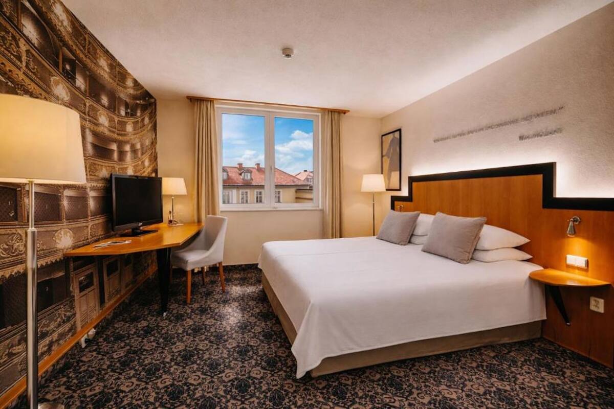 A hotel room with a bed, desk, TV, and a view of buildings through the window.