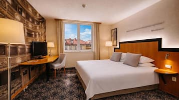 A hotel room with a bed, desk, TV, and a view of buildings through the window.