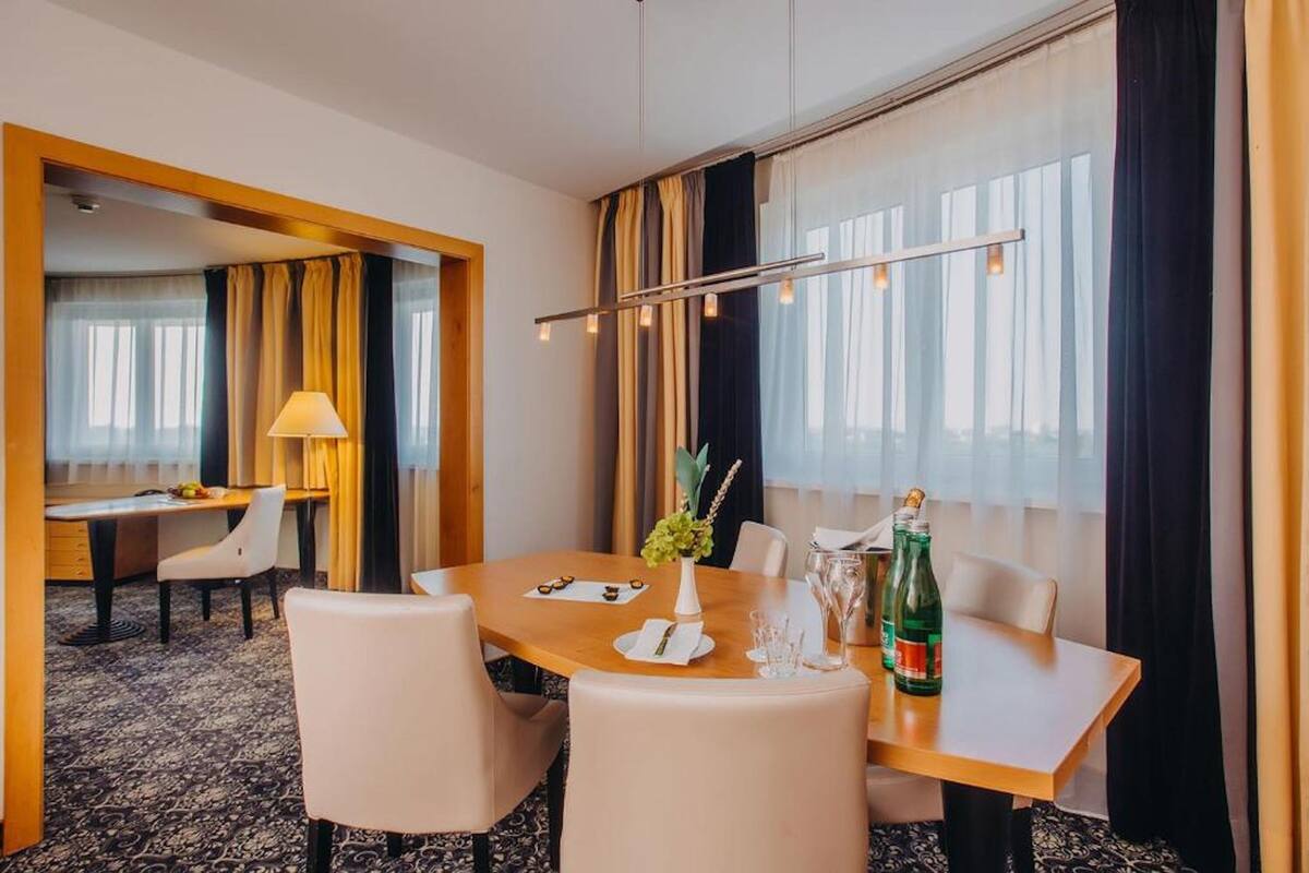 A hotel room with a dining area featuring a wooden table, white chairs, a vase with flowers, and a bottle of champagne.