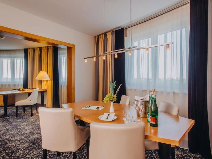 Habitación de hotel con zona de comedor que incluye una mesa de madera, sillas blancas, un florero con flores y una botella de champán.