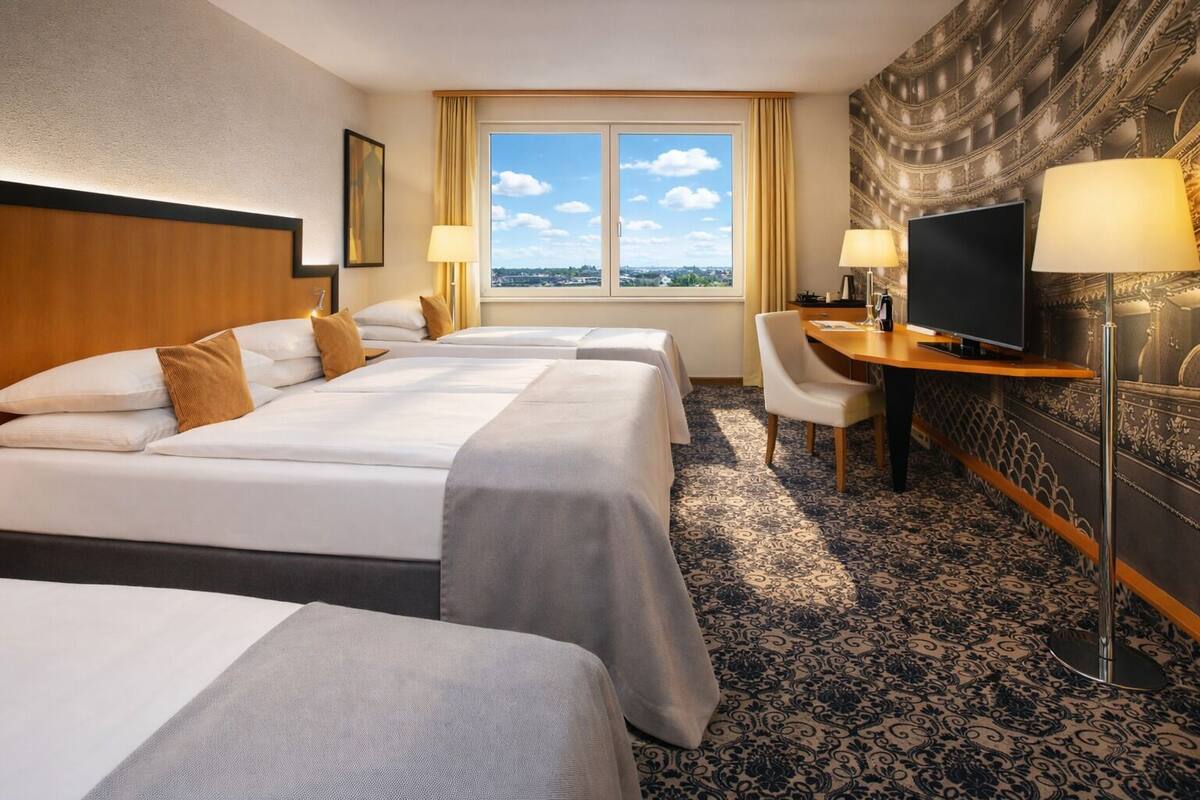 A hotel room with three beds, a desk with a TV, and a large window with a city view.