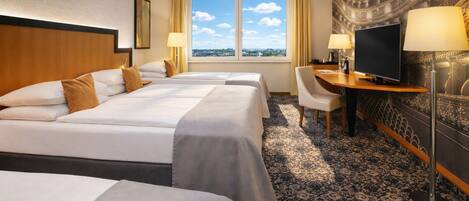 A hotel room with three beds, a desk with a TV, and a large window with a city view.