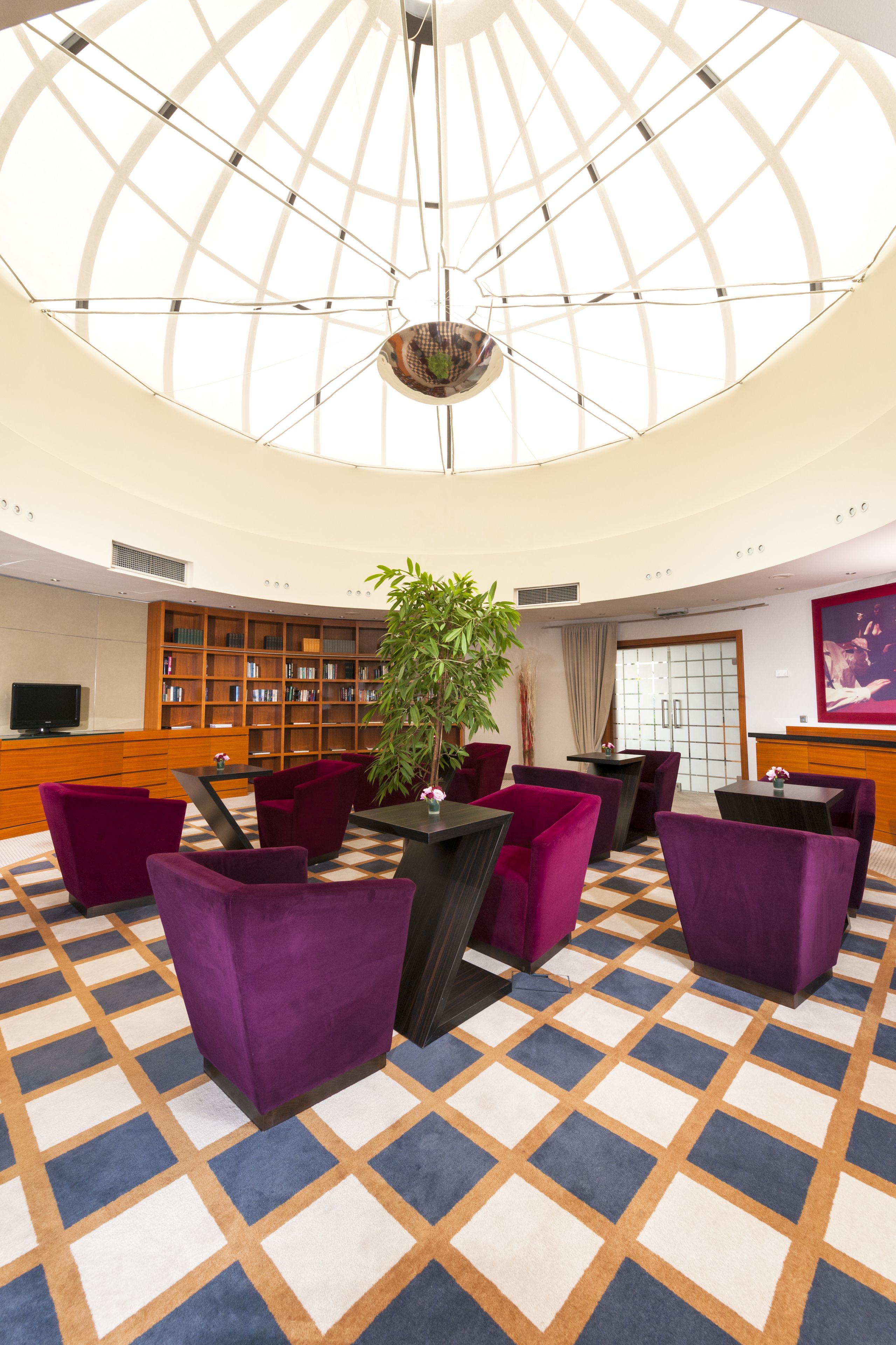 A lobby with a high ceiling, a central plant, purple seating, a bookshelf, and a television.