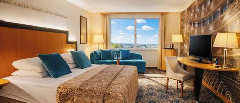 A hotel room with a bed, two sofas, a desk with a TV, and a large window with a city view.