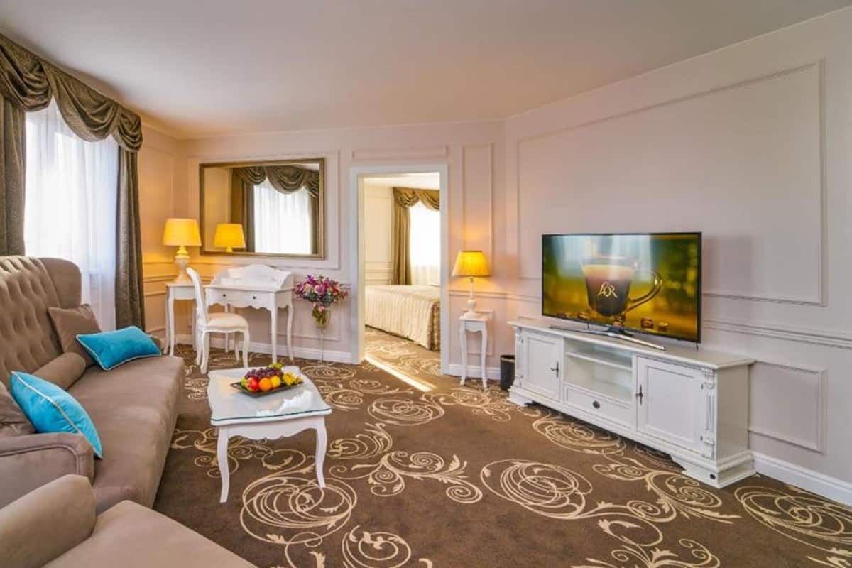 A hotel room with a brown sofa, a dining table with chairs, a TV, and a bed in the background.