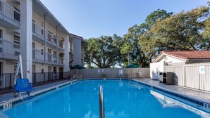 A swimming pool with a metal railing in the foreground, surrounded by a building with multiple balconies and a covered area with umbrellas.