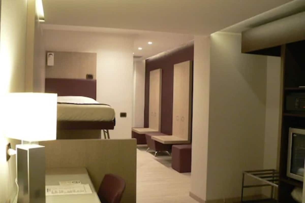 A hotel room with a bed, a desk with a chair, a TV, and a seating area with a bench.