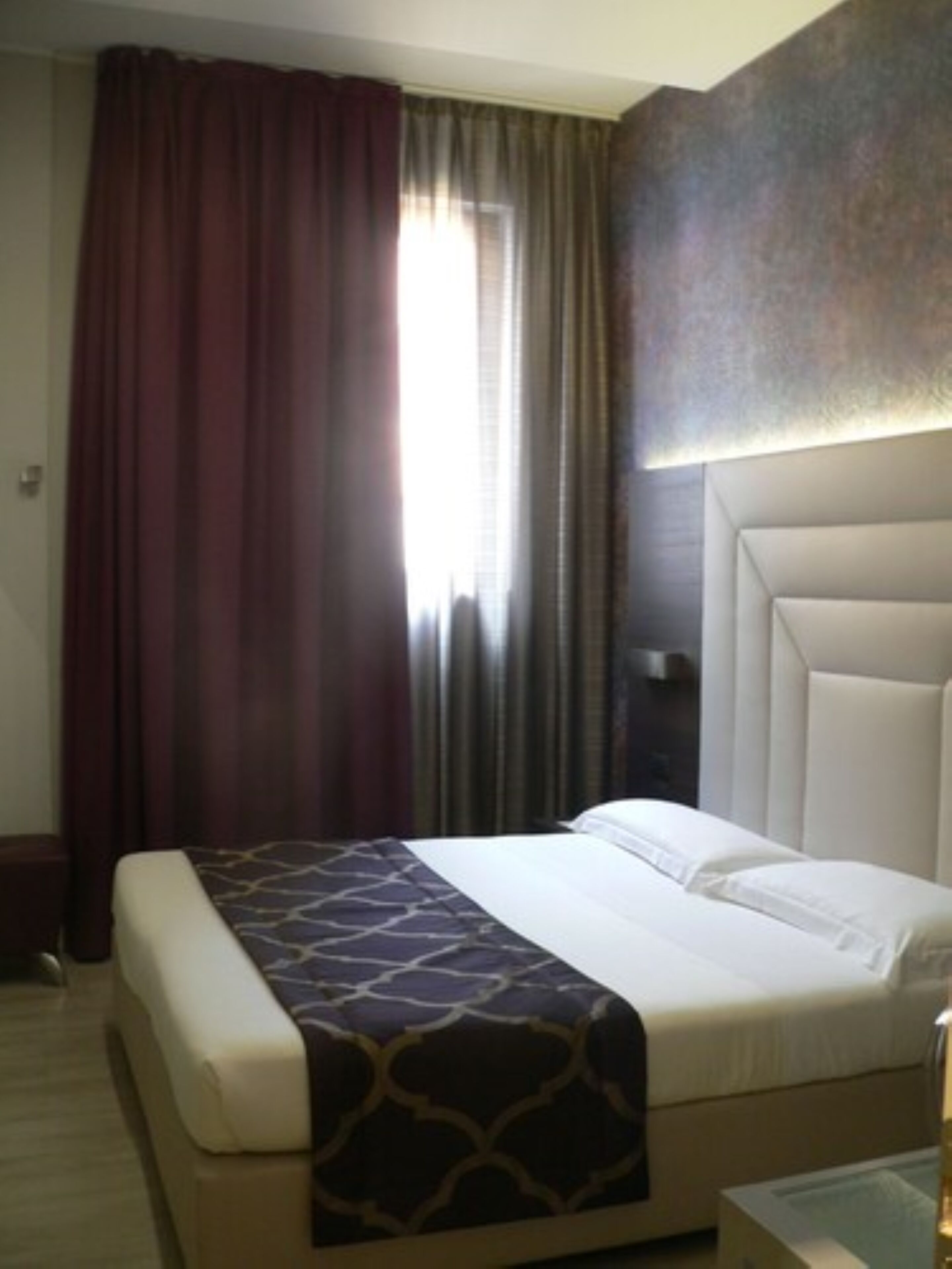 A hotel room with a large bed, a headboard, a bedside table, and a window with curtains.