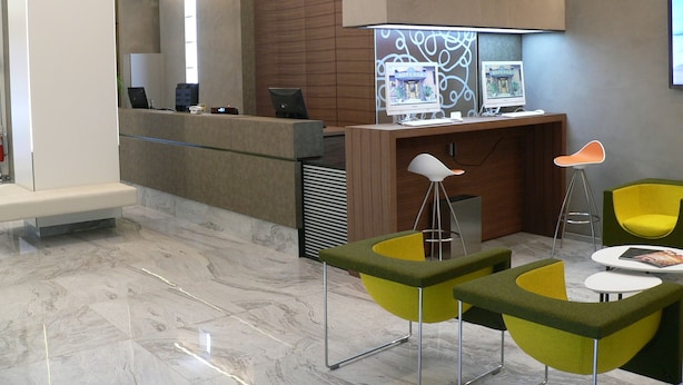 Un área de recepción con mostrador, dos monitores de ordenador, una silla verde y amarilla, una silla blanca y una pequeña mesa con una revista.