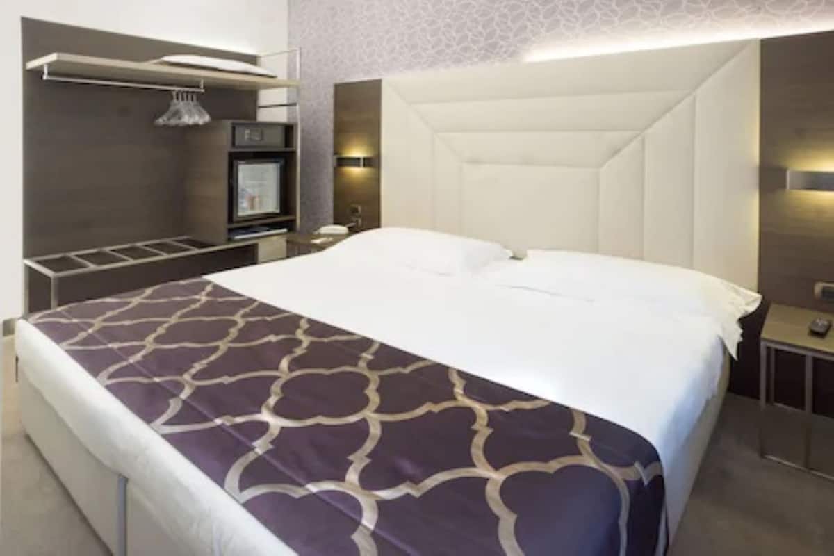 A modern hotel room with a large bed, a built-in TV, a minibar, and a seating area with a table and chairs.