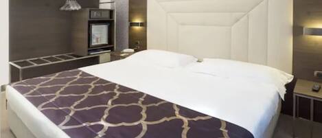 A modern hotel room with a large bed, a built-in TV, a minibar, and a seating area with a table and chairs.