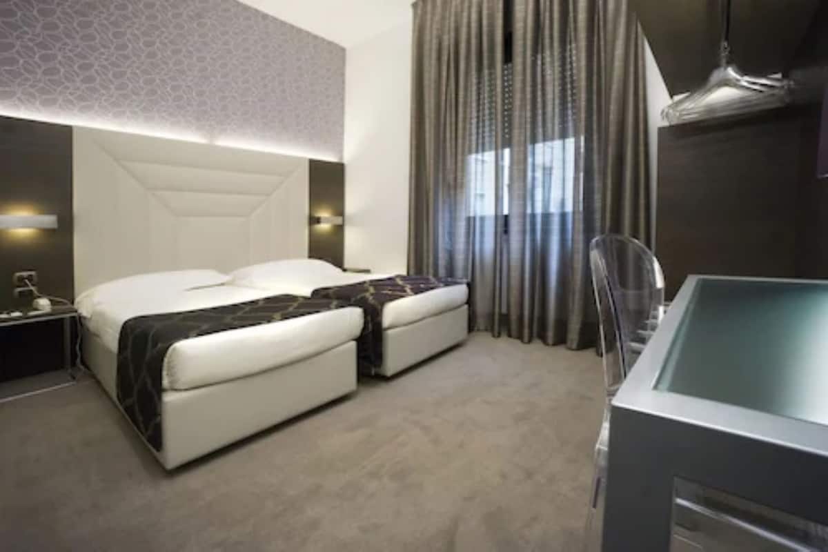 A modern hotel room with a large bed, a separate sofa, a glass-enclosed bathtub, and a large window with curtains.