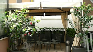 Un área de patio al aire libre con mesas y sillas, rodeada de plantas en macetas y una pérgola.