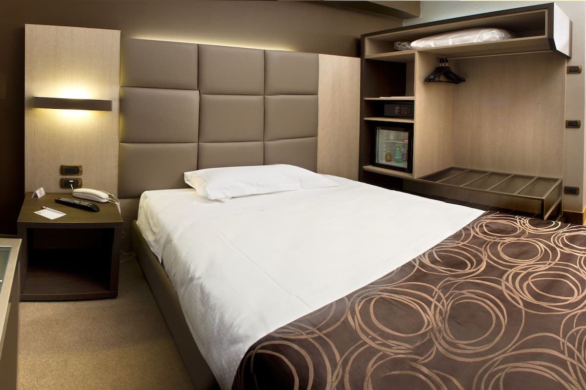 A hotel room with a bed, a bedside table with a phone and a notepad, a headboard, a shelf with a bottle and a remote control, and a small cabinet with a mirror and a light.