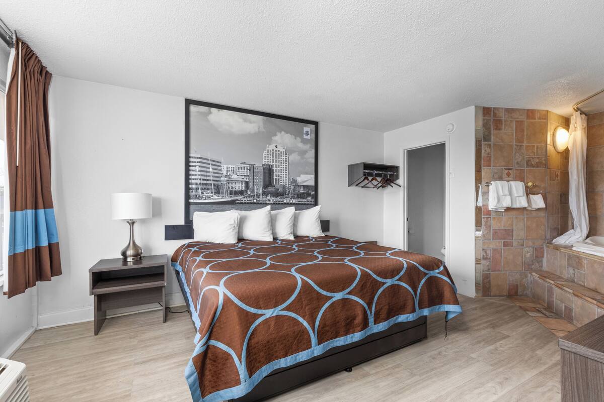Une chambre d’hôtel comprenant un lit, une table de chevet, une lampe, une salle de bain avec douche et une œuvre d’art représentant un paysage urbain accrochée au mur.