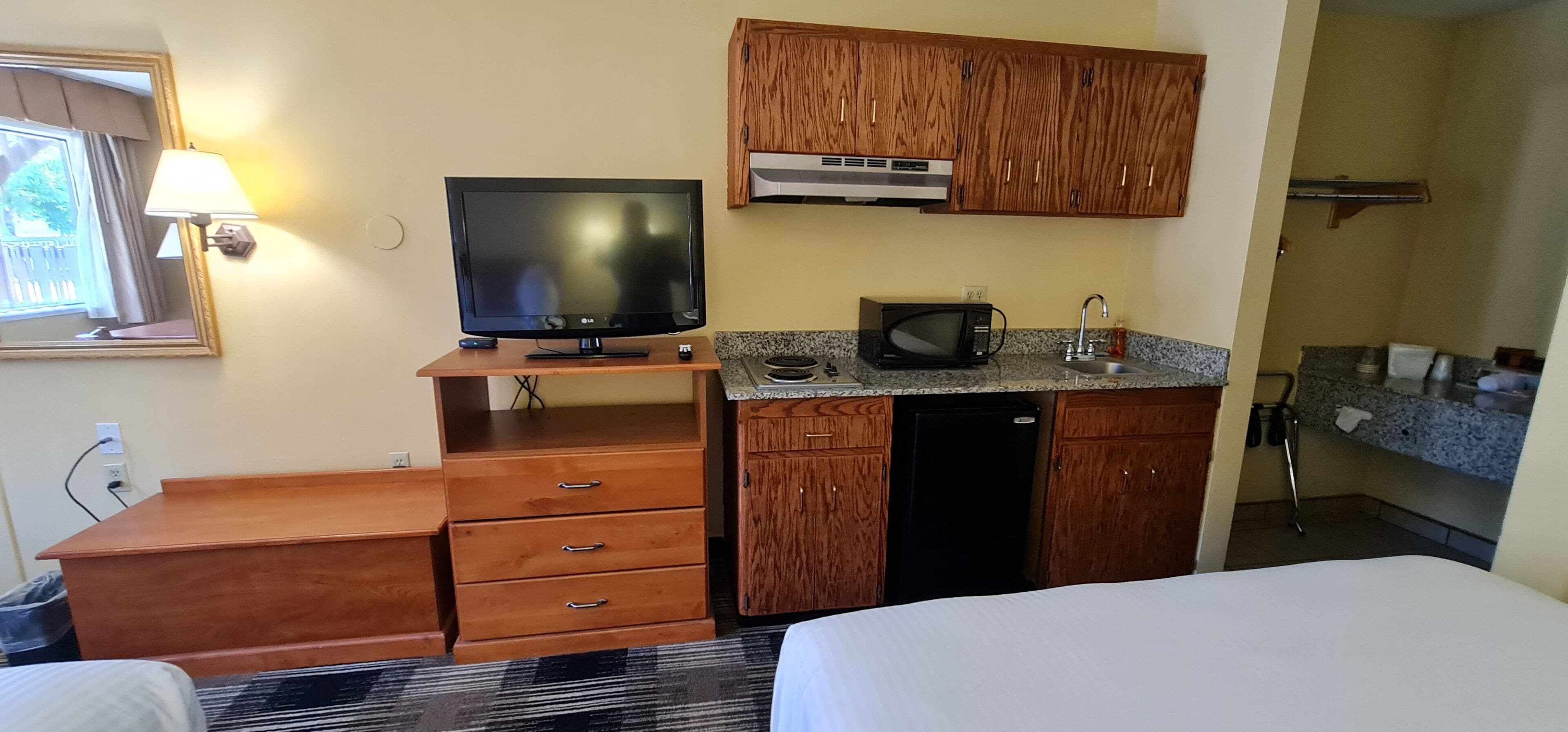 Una habitación de hotel con cama, televisor sobre un mueble de madera, kitchenette con microondas y lavabo, y un armario de madera sobre la encimera.