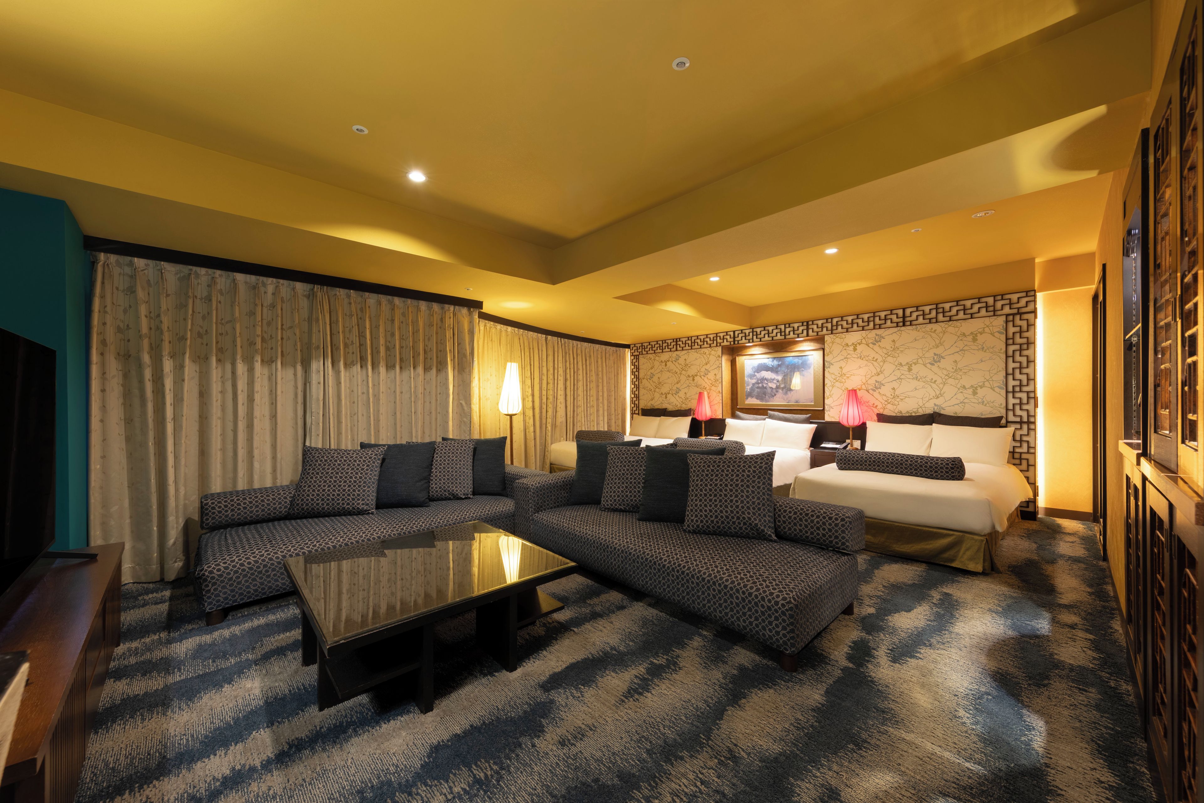 A hotel room with a sectional sofa, a bed, a flat-screen TV, and a patterned carpet.