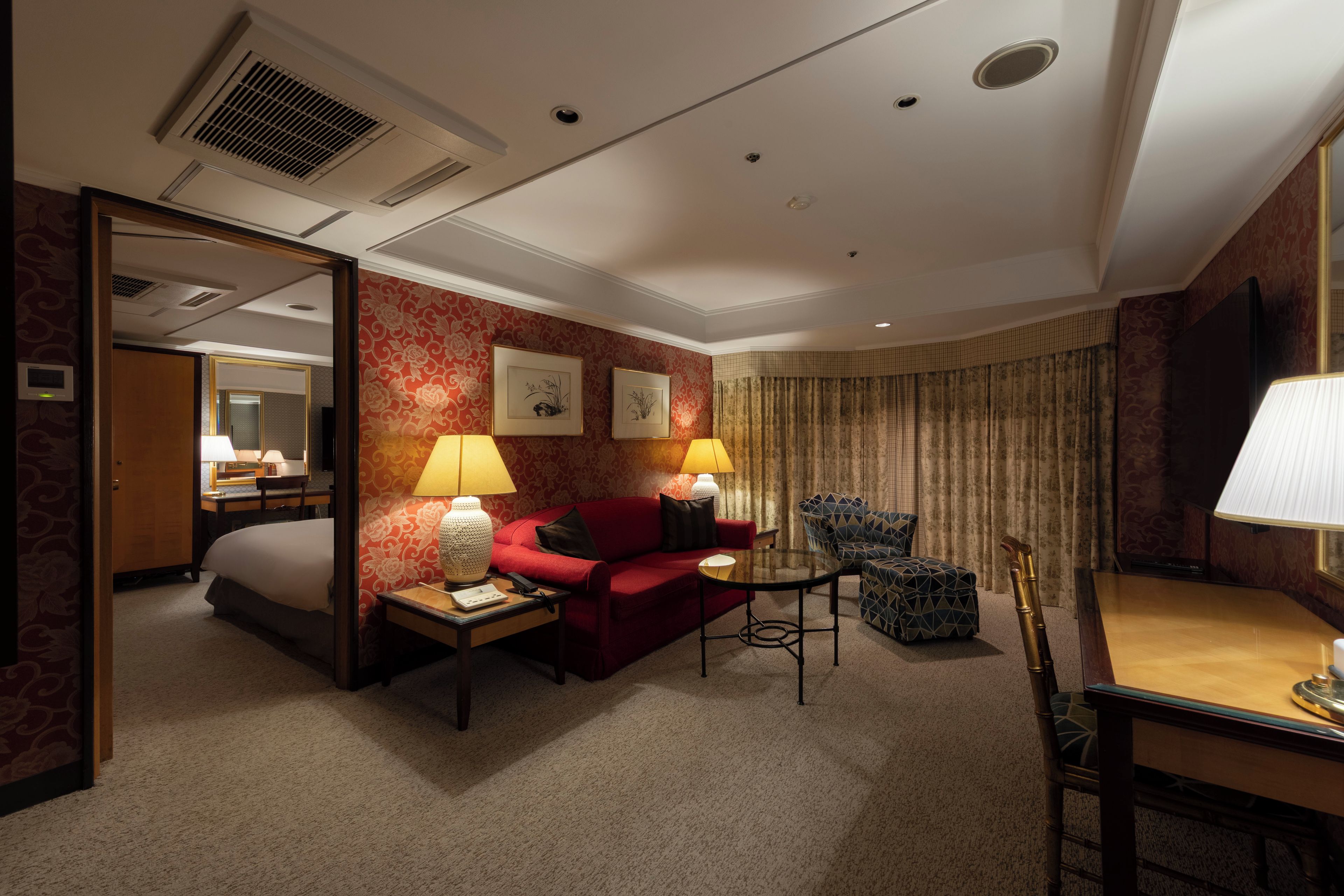 A hotel room with a bed, a red sofa, a small table with lamps, a chair, and a desk with a lamp.