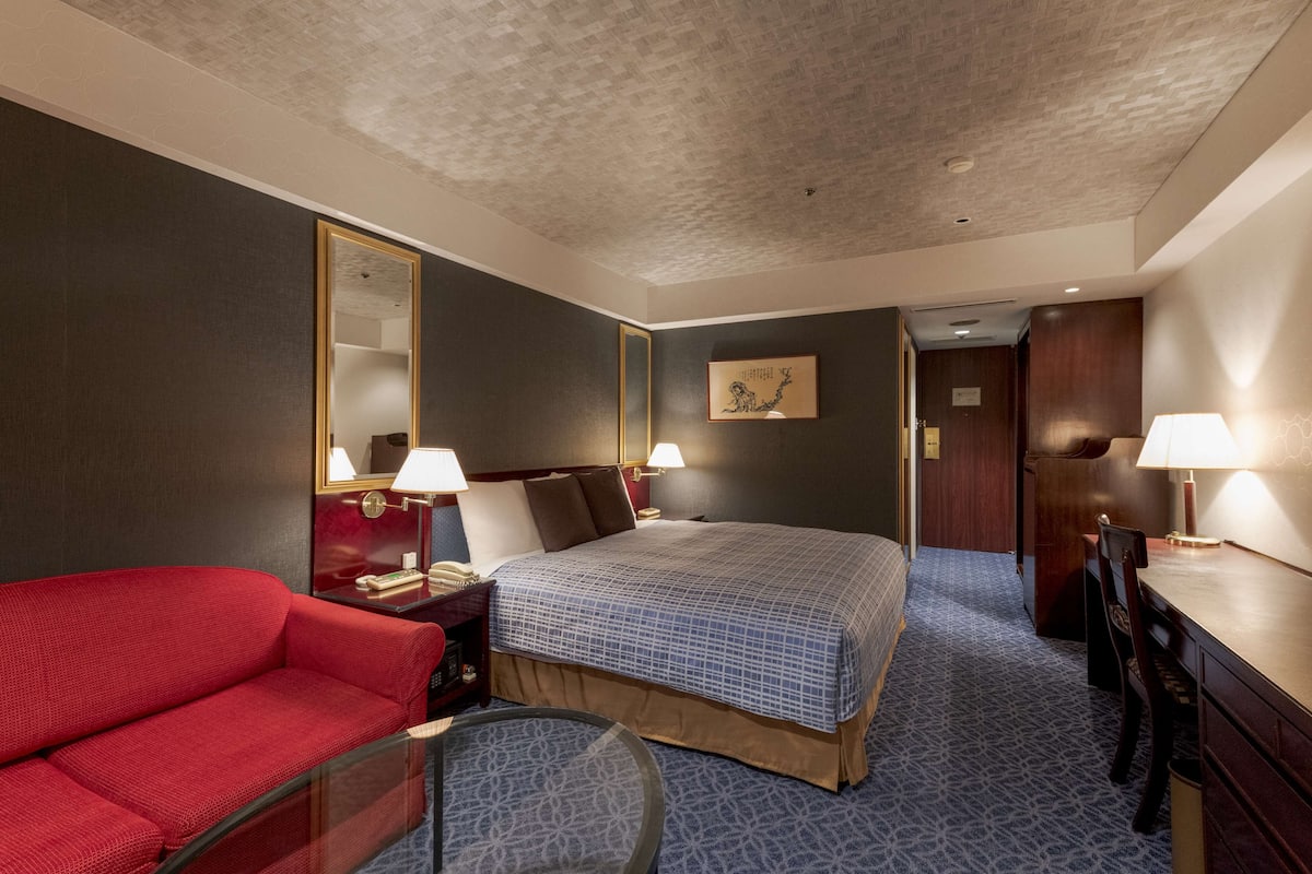 Habitació d'hotel amb un llit gran, un sofà vermell, una taula de nit amb làmpada i un mirall.