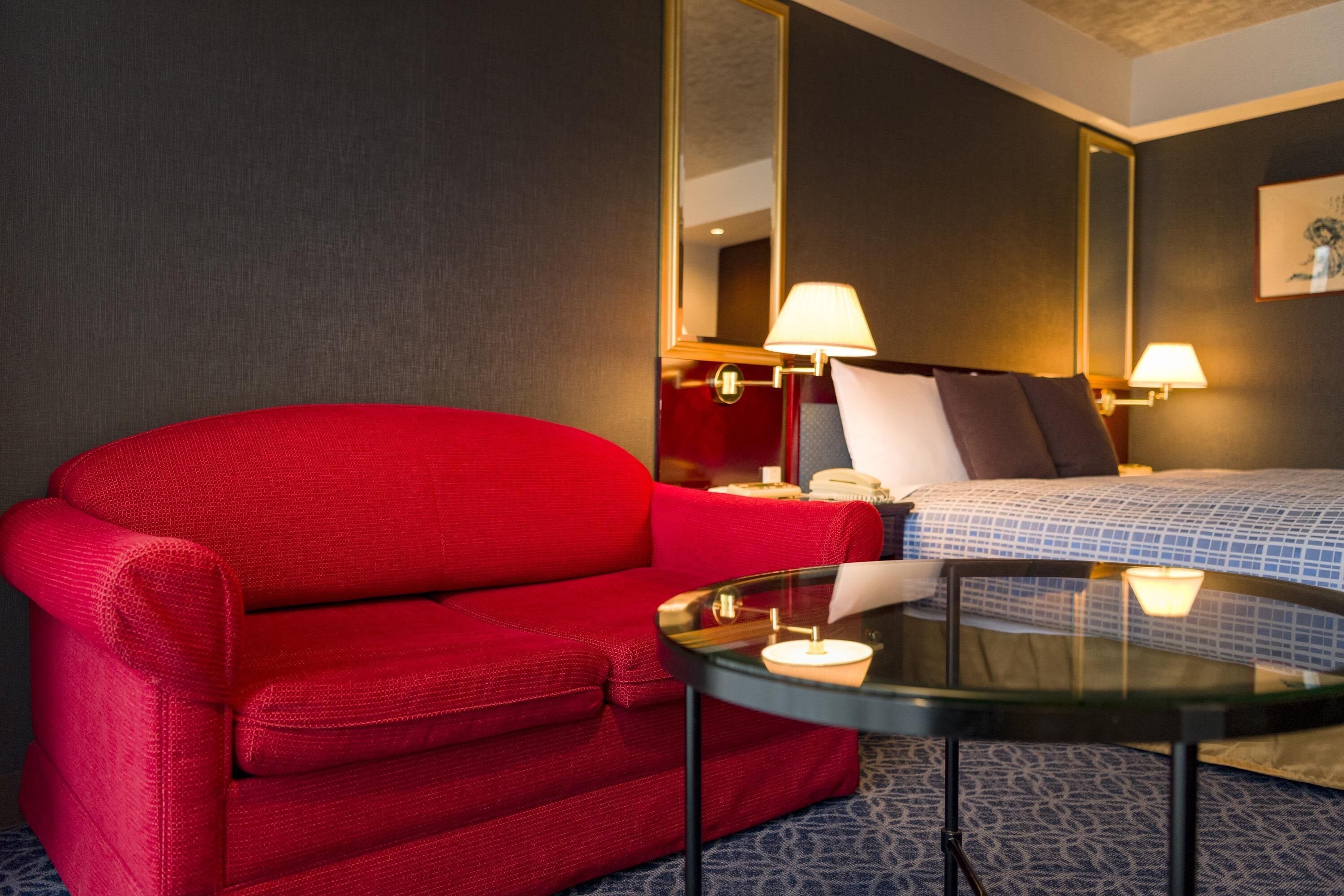 A hotel room with a red sofa, a round glass table, a bed with a blue and white checkered bedspread, and two wall-mounted lamps.