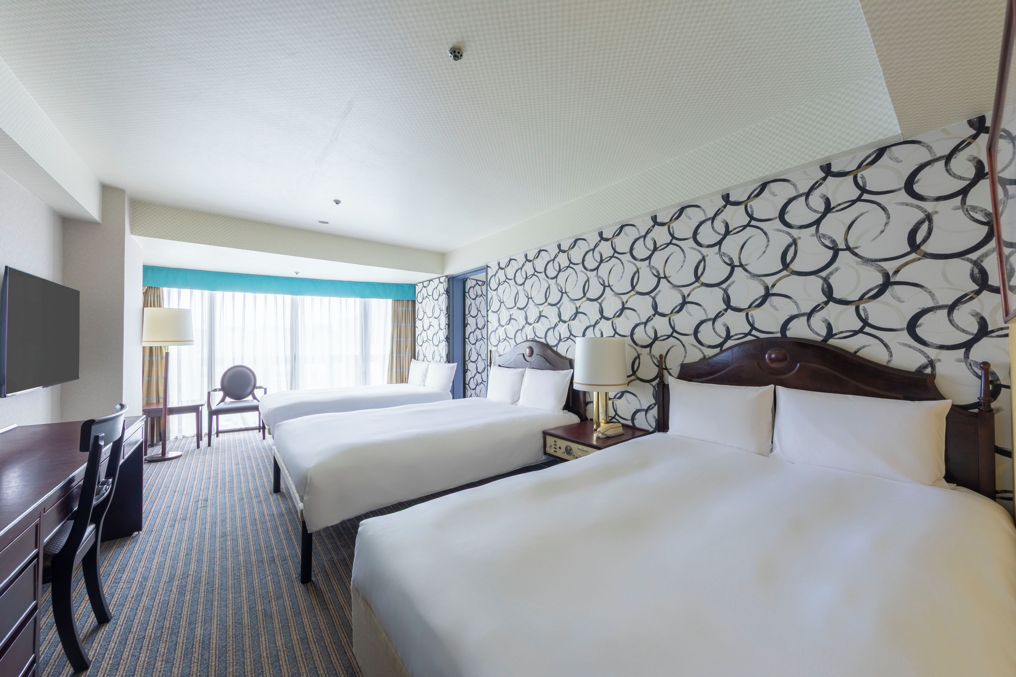 A hotel room with two beds, a desk, a chair, a TV, and a patterned wall.