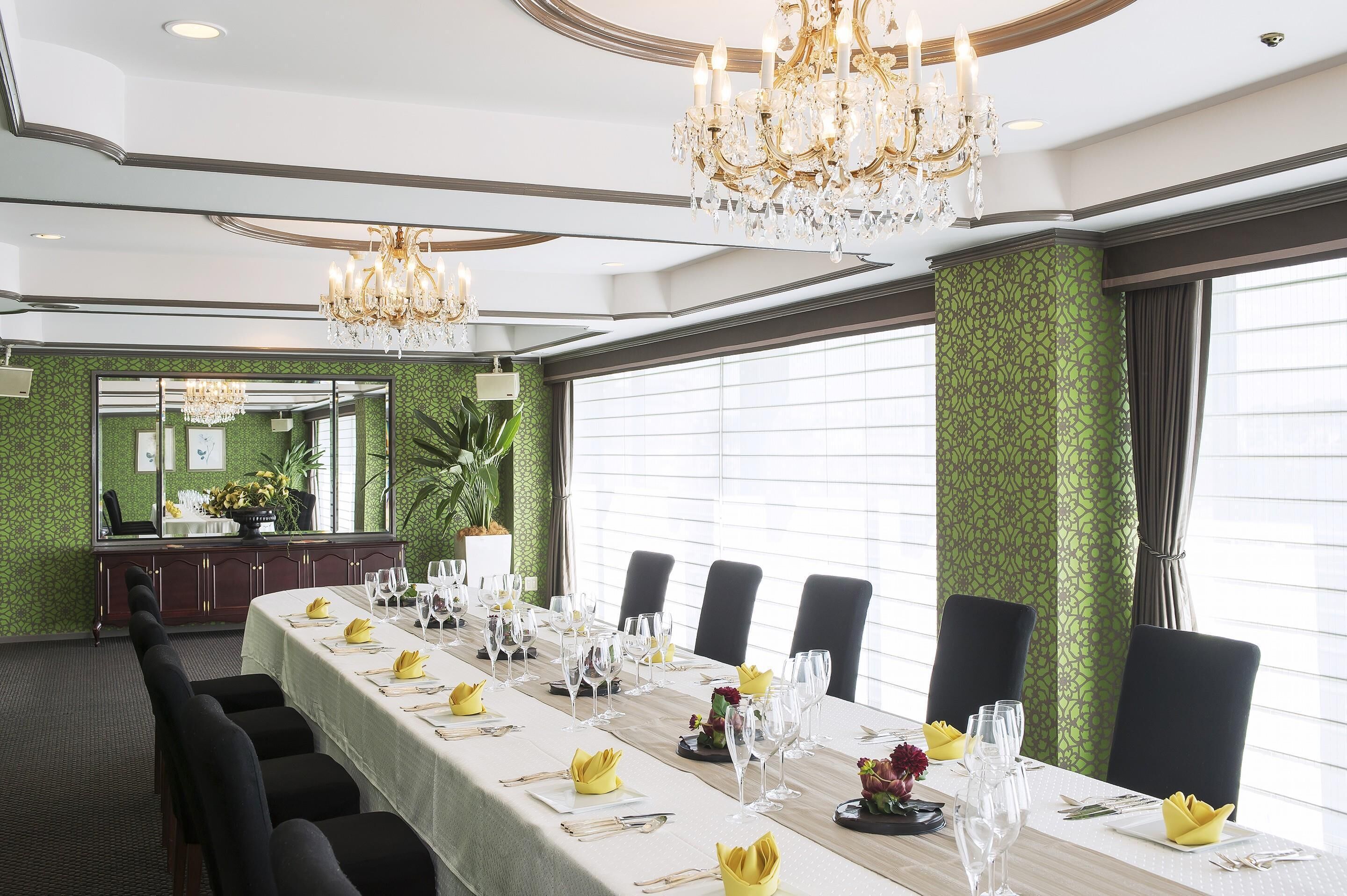 A formal dining room with a long table set for a meal, chandeliers, and a green patterned wall.