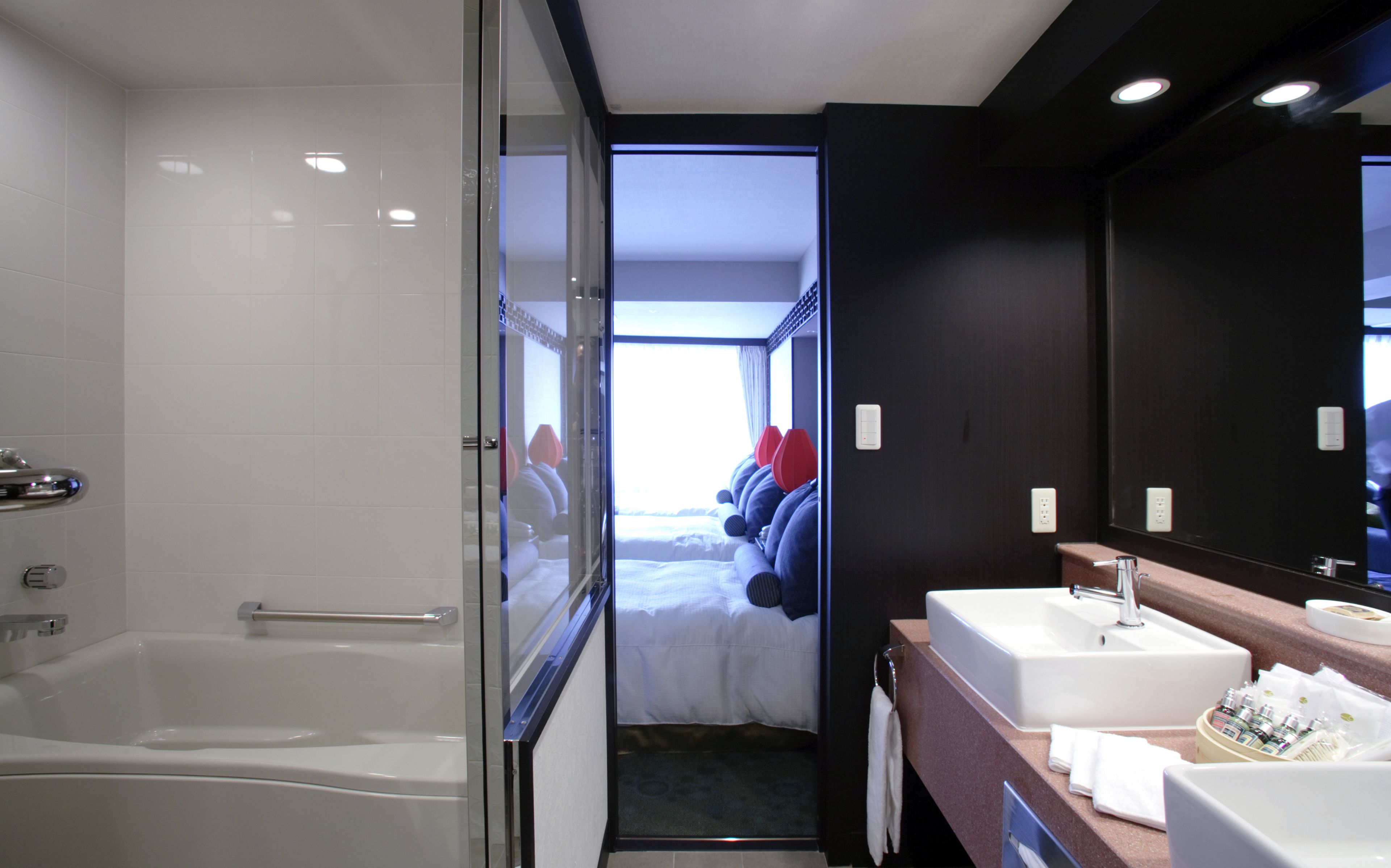 A modern bathroom with a glass-enclosed shower, a large bathtub, a vanity with a mirror, and a bed visible through an open door.