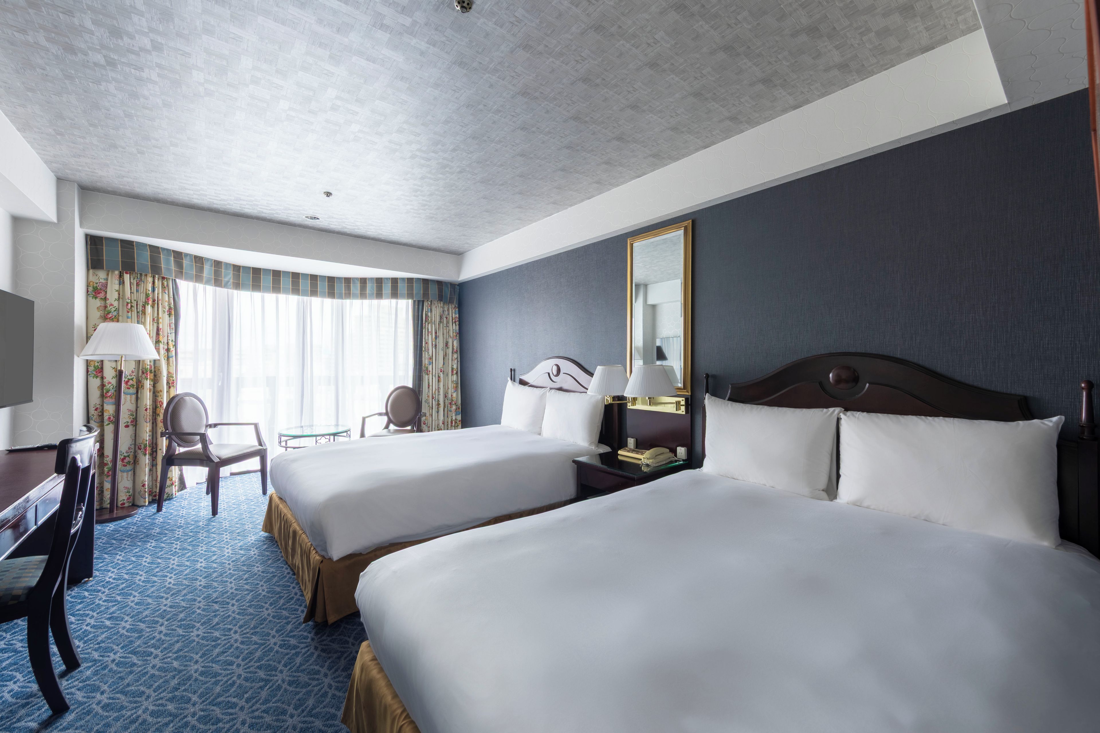 A hotel room with two beds, a desk, a chair, a mirror, and a window with curtains.