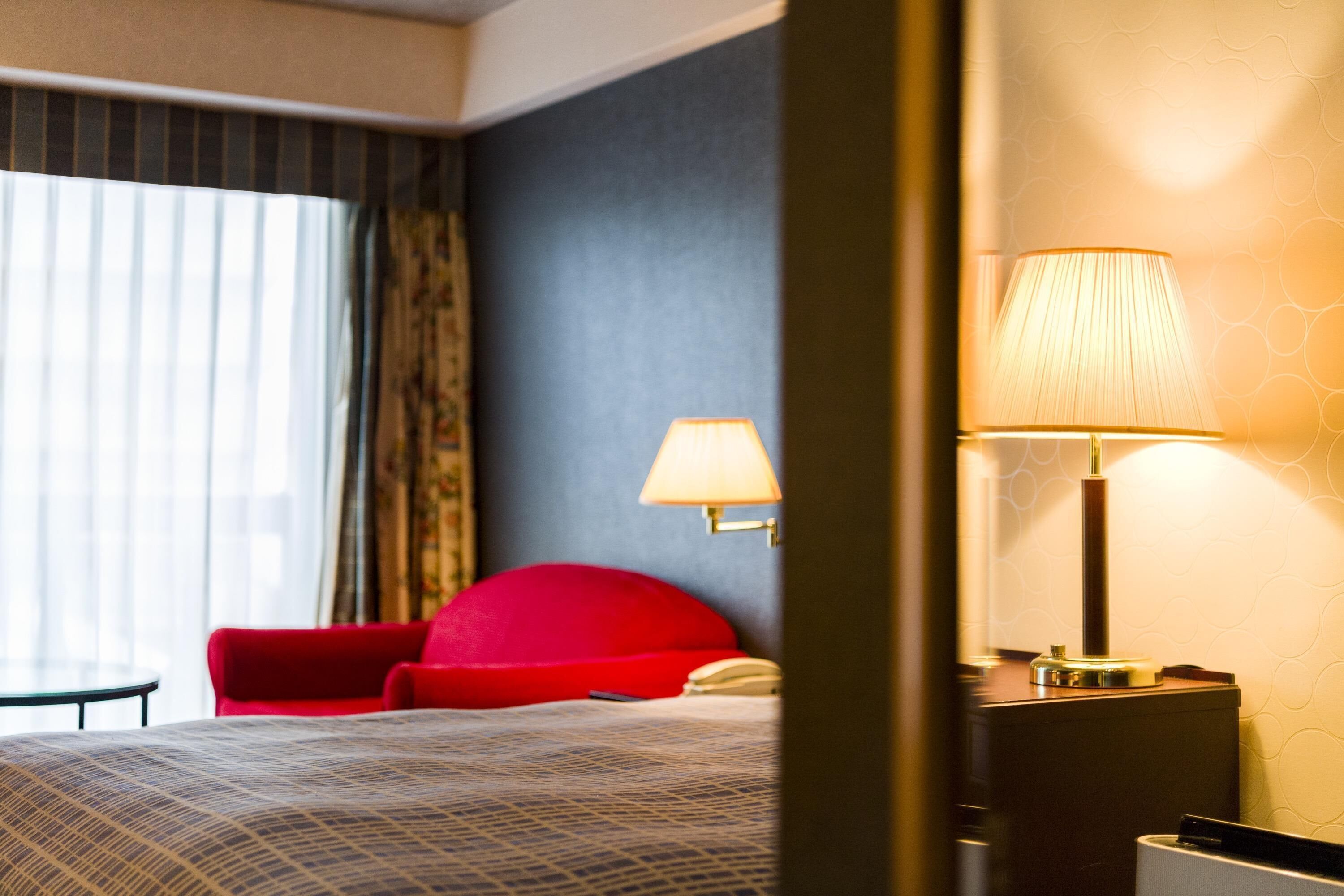 A hotel room with a bed, a red chair, a lamp, and a window with curtains.