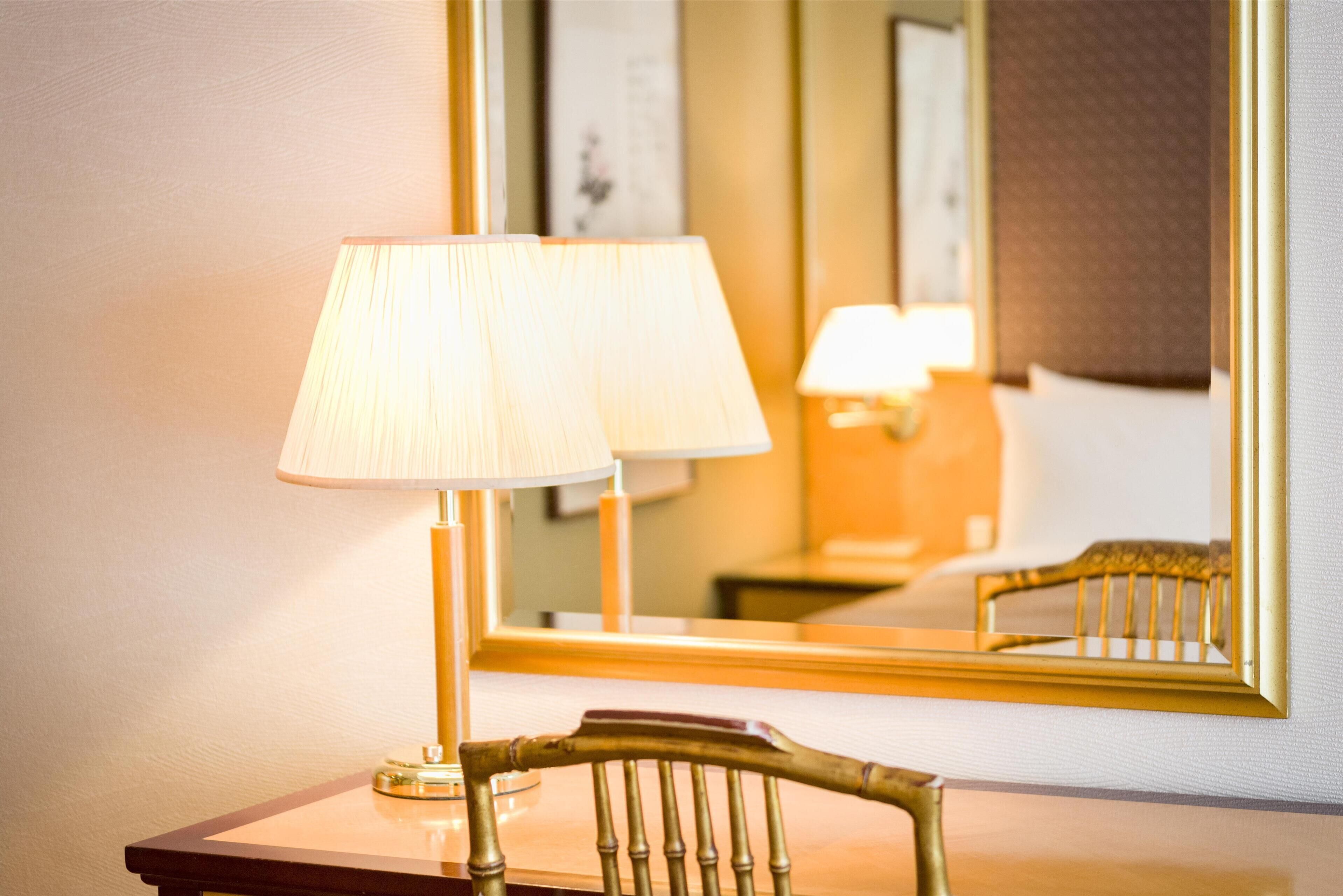 Habitació d'hotel amb una cadira de fusta, un taulell i una làmpada. Un mirall ampli reflecteix l'interior de l'habitació.