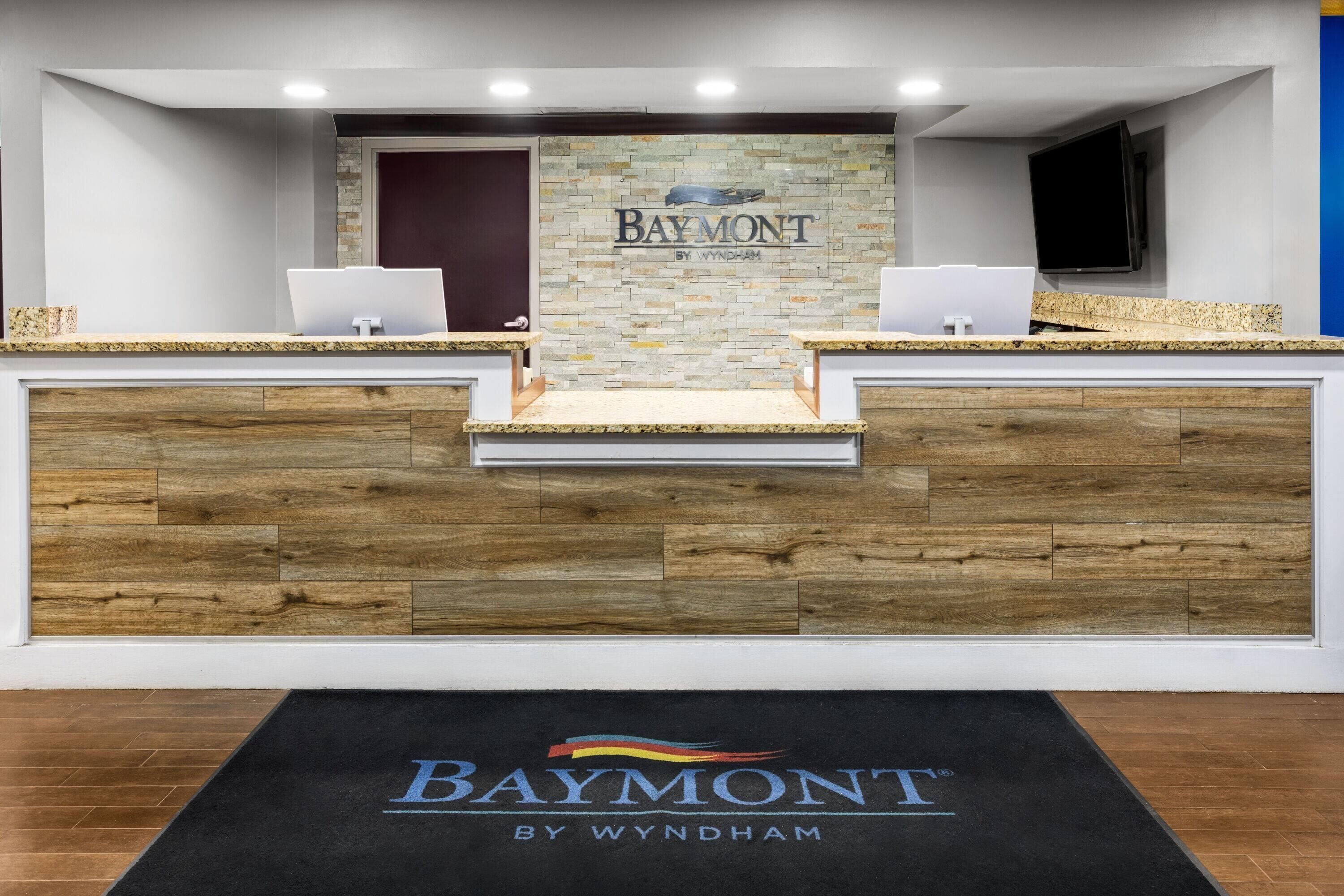 A reception desk with a stone wall backdrop and a black mat with 'Baymont by Wyndham' branding.