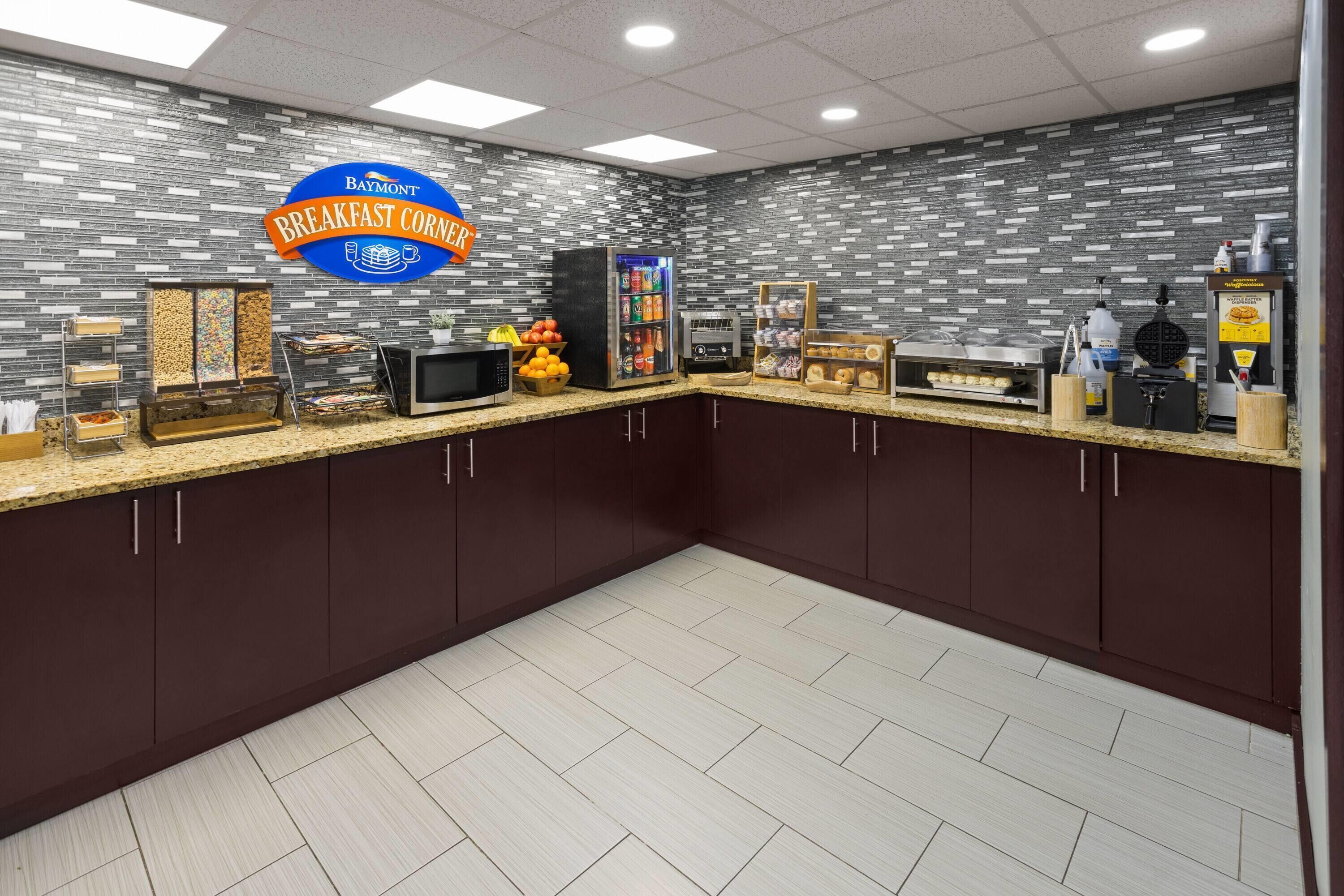 Rincón de desayuno con cartel de Baymont, microondas, exhibición de frutas, máquina para hacer gofres y dispensador de masa para gofres.