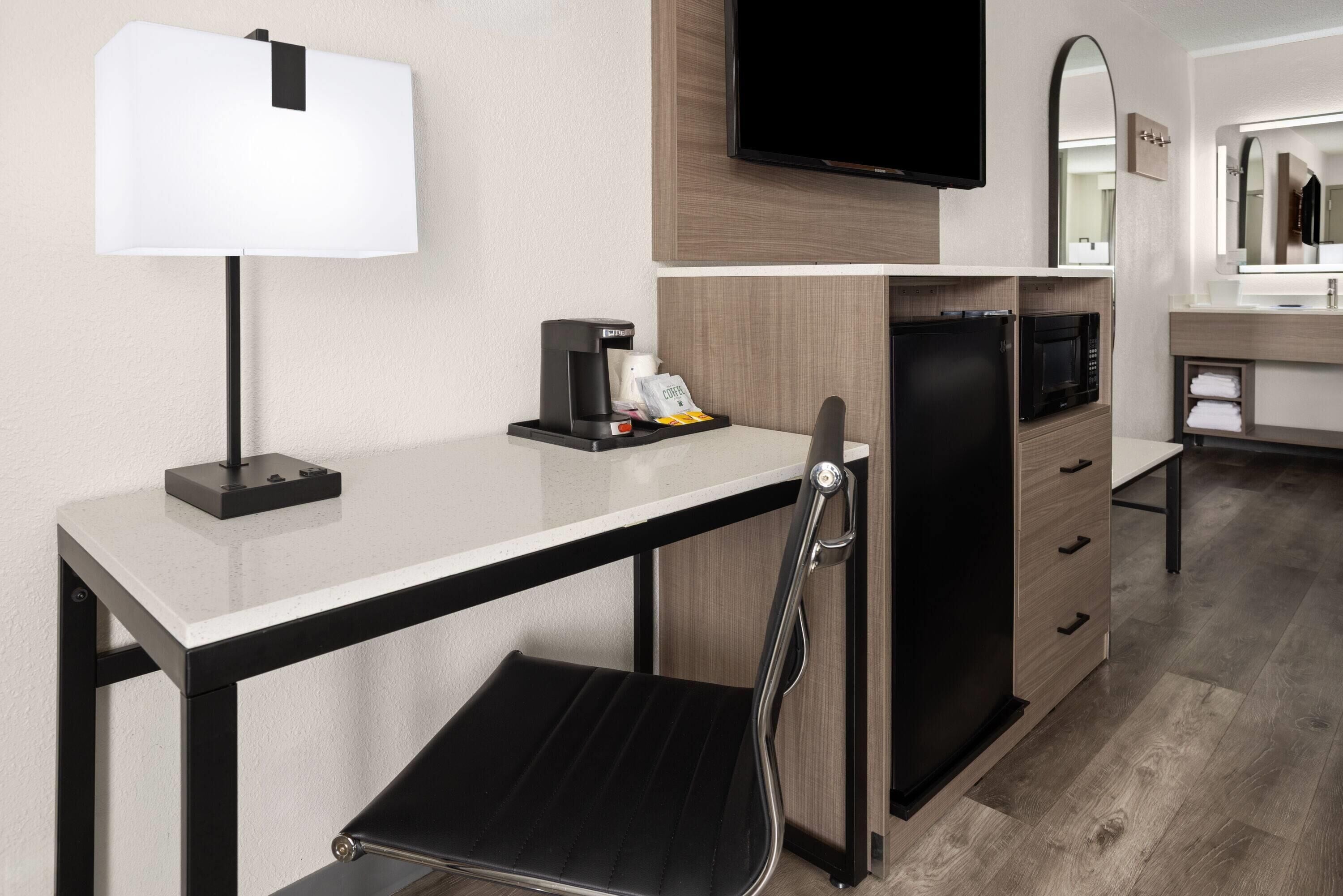 A hotel room with a desk, a chair, a flat-screen TV, a coffee maker, a microwave, and a mirror.