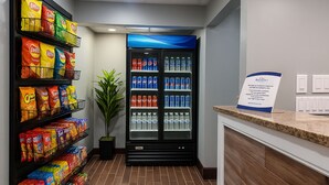 A hotel lobby with snack shelves, a beverage refrigerator, a reception desk, and a potted plant.