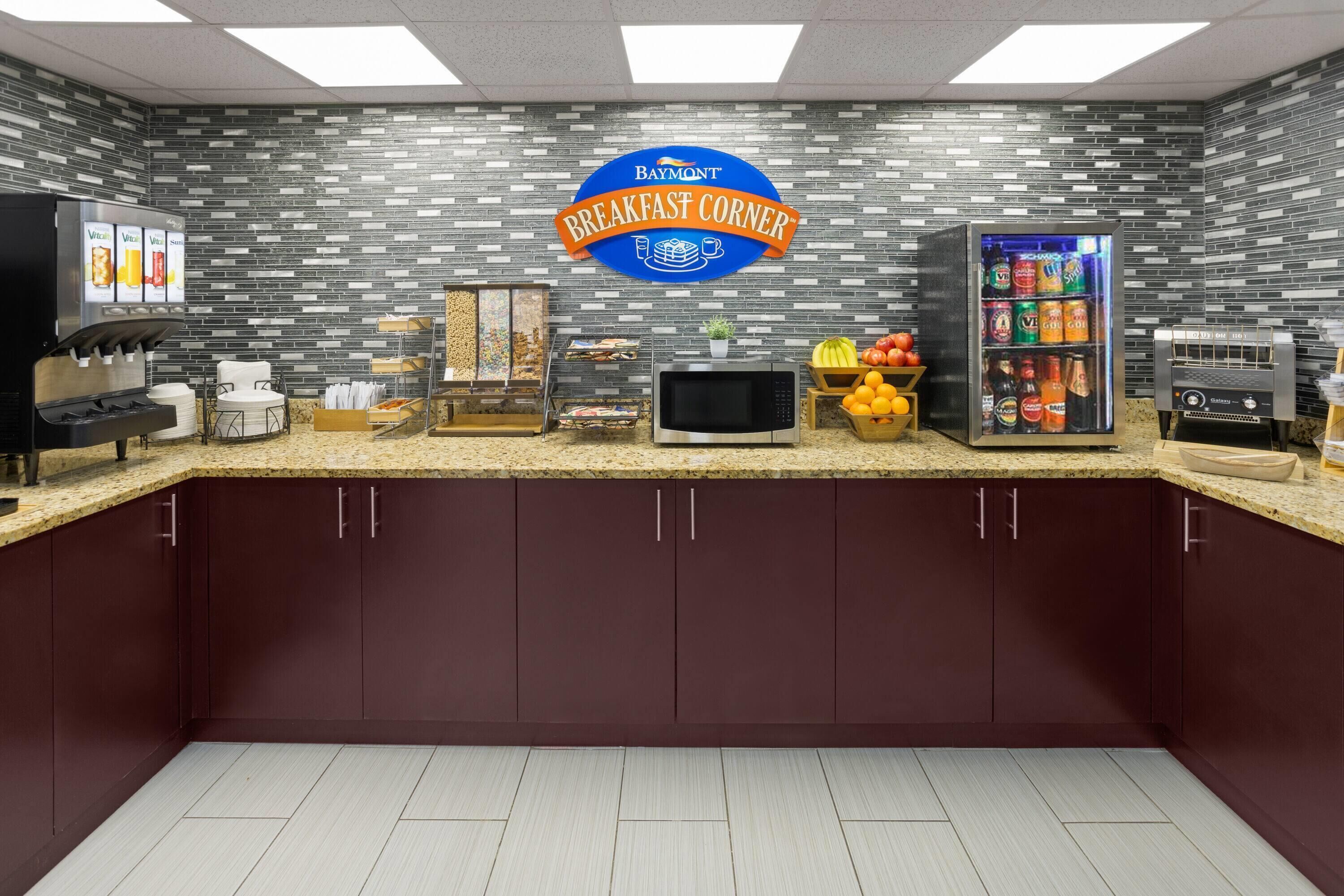 A breakfast area with a sign reading 'Breakfast Corner,' a beverage dispenser, a microwave, a fruit display, and a refrigerator.