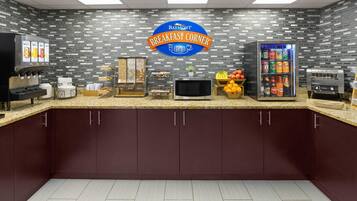 A breakfast area with a sign reading 'Breakfast Corner,' a beverage dispenser, a microwave, a fruit display, and a refrigerator.